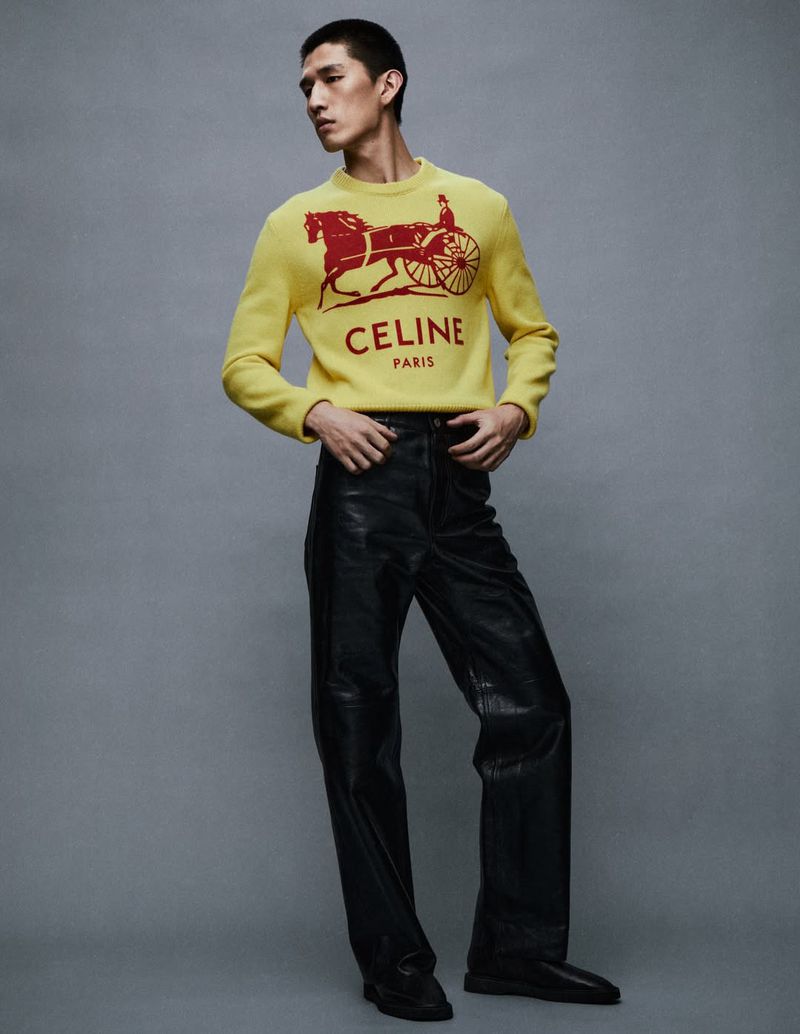 A young Asian man poses indoors wearing a yellow Celine sweater with a horse and carriage logo and black leather pants, against a grey backdrop.