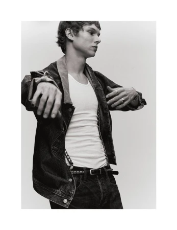 Monochrome portrait of a young adult male in a denim jacket and white t-shirt against a plain background in a studio setting.