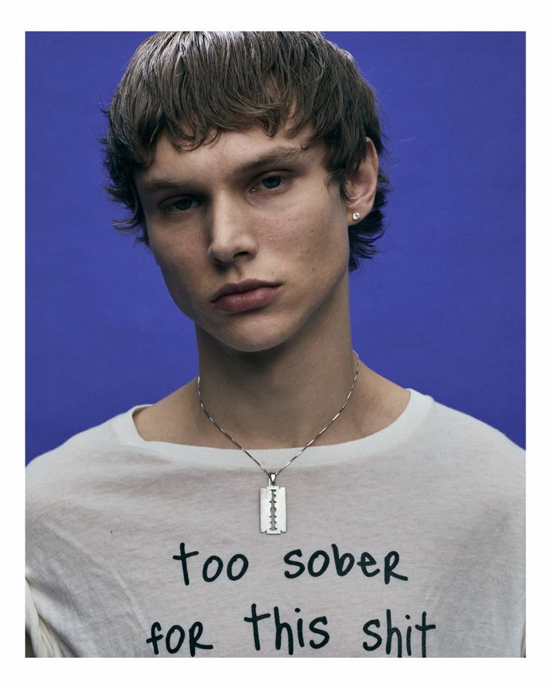 Portrait of a young adult man with fair skin and light brown hair wearing a white t-shirt with a silver razor blade necklace on a blue background.