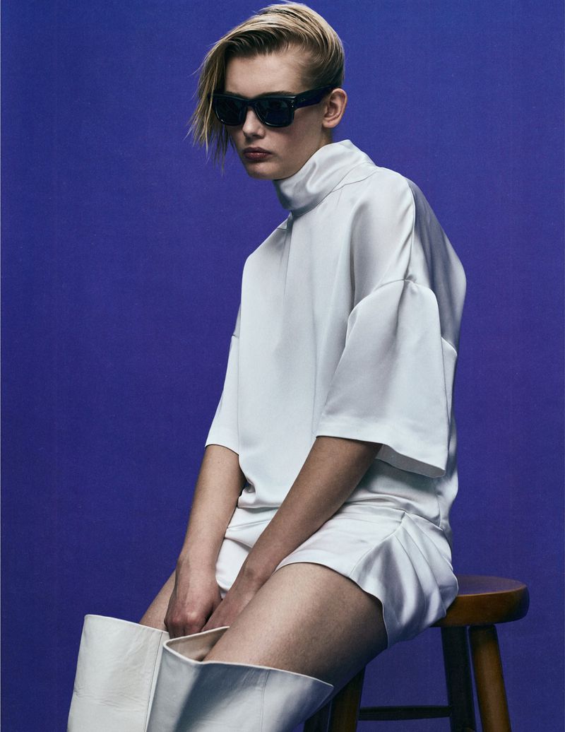 Fashion portrait of a blond adult wearing sunglasses and an all-white outfit, seated on a stool against a solid violet backdrop.
