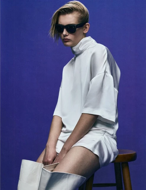 Fashion portrait of a blond adult wearing sunglasses and an all-white outfit, seated on a stool against a solid violet backdrop.