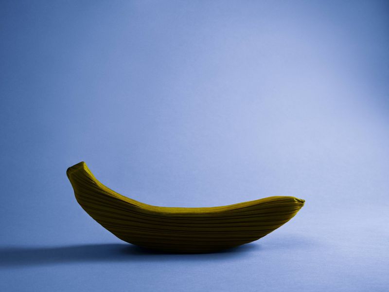 Studio shot of a yellow banana sculpture made of corrugated material against a light blue background, emphasizing texture and minimalist art.