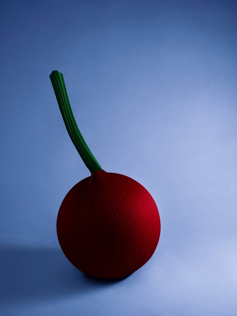 Stylized image of a red, spherical fruit or vegetable with a green stem on a blue gradient background, creating a surreal, minimalist vibe.