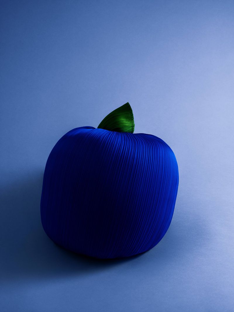 Still life of a dark blue rounded object with a green leaf, set against a soft blue gradient background, in a studio setting.