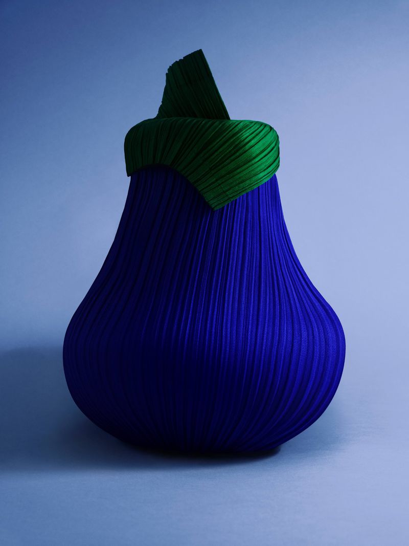 An abstract eggplant-shaped sculpture with a vertically ribbed blue body and a layered green stem stands against a light blue background.