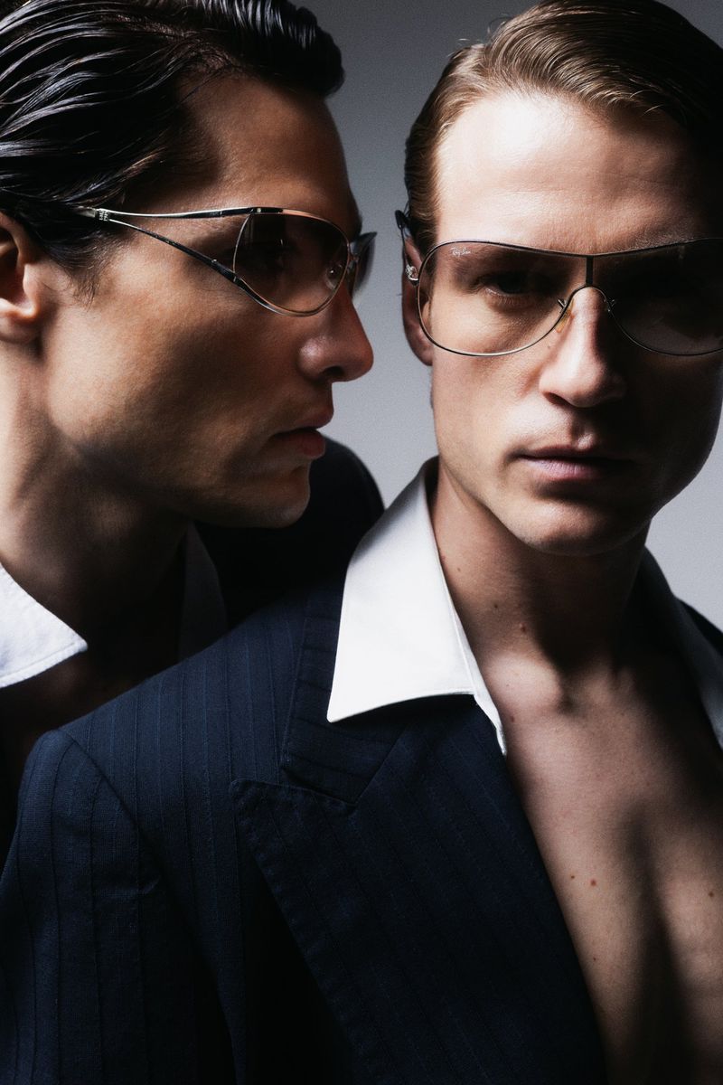 Portrait of two adult males in suits and sunglasses posing in front of a plain studio backdrop with diffused lighting and dark coloration.