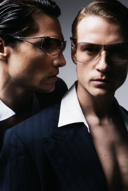 Portrait of two adult males in suits and sunglasses posing in front of a plain studio backdrop with diffused lighting and dark coloration.