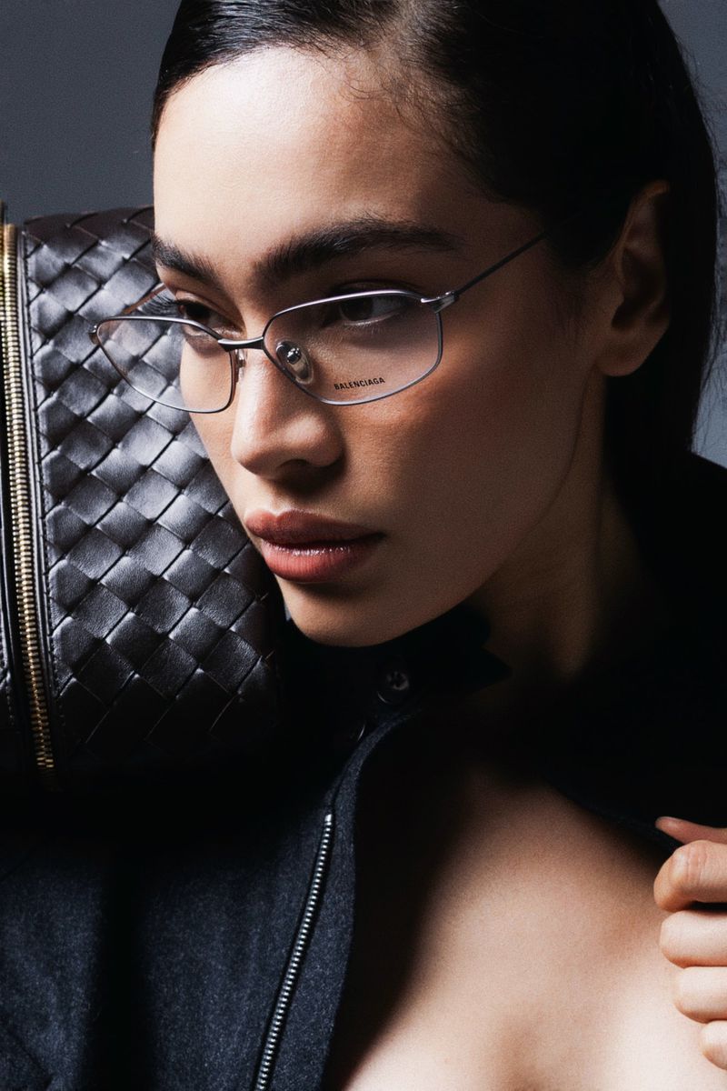 Portrait of a woman with glasses holding a large woven leather bag over her shoulder in a close up shot in a photo studio.