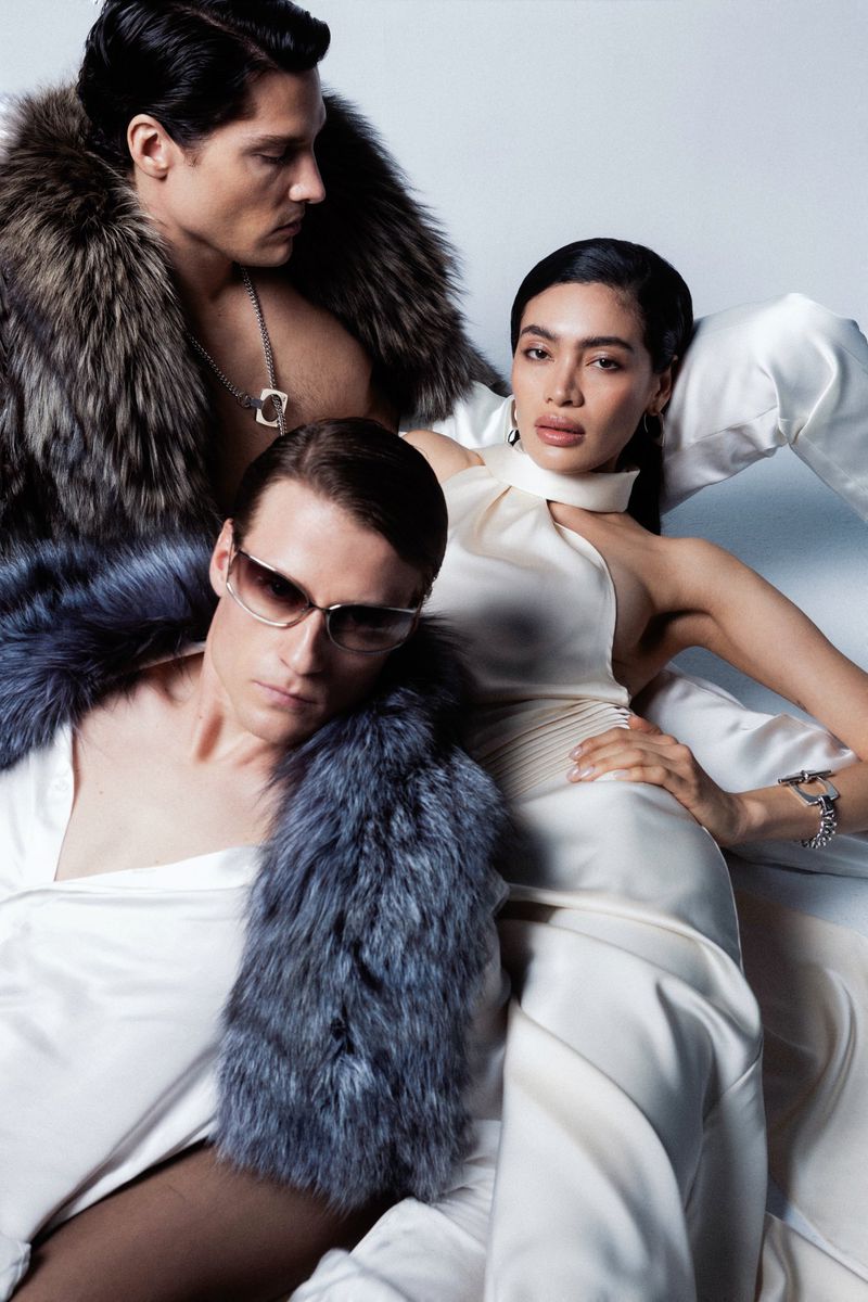 Fashion shot featuring an Asian woman in a cream-colored dress with two men, one in sunglasses, all wearing fur shawls, indoors.