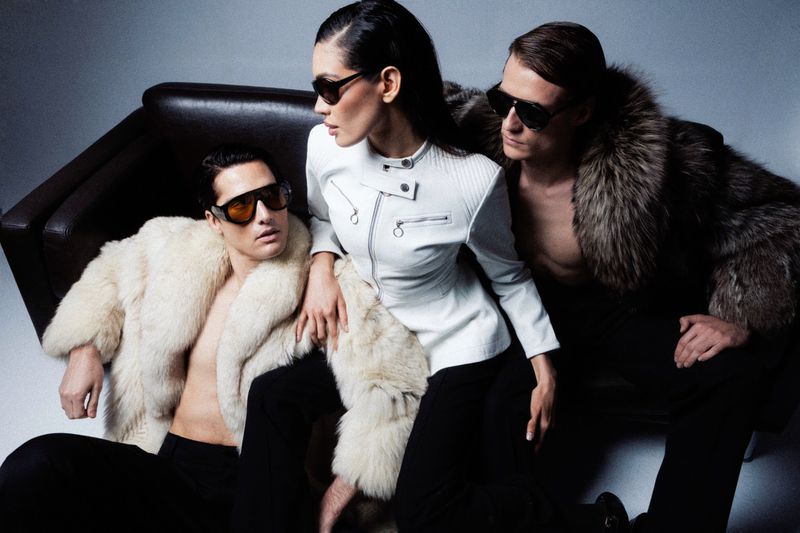 Fashion shot of three adults in sunglasses and fur coats posing on a dark couch in a studio. One woman and two men are featured.