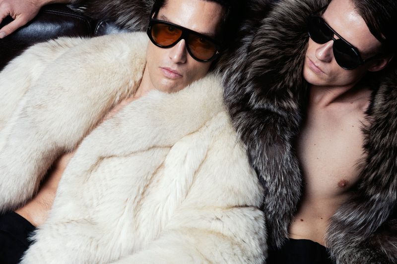 Two adult men wearing sunglasses and fur coats, lying down indoors with low-key lighting, creating a fashionable and luxurious aesthetic.