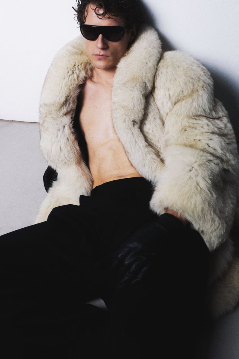 An adult male with sunglasses poses in an off-white fur coat with black trousers and gloves, looking at the camera in a studio setting.