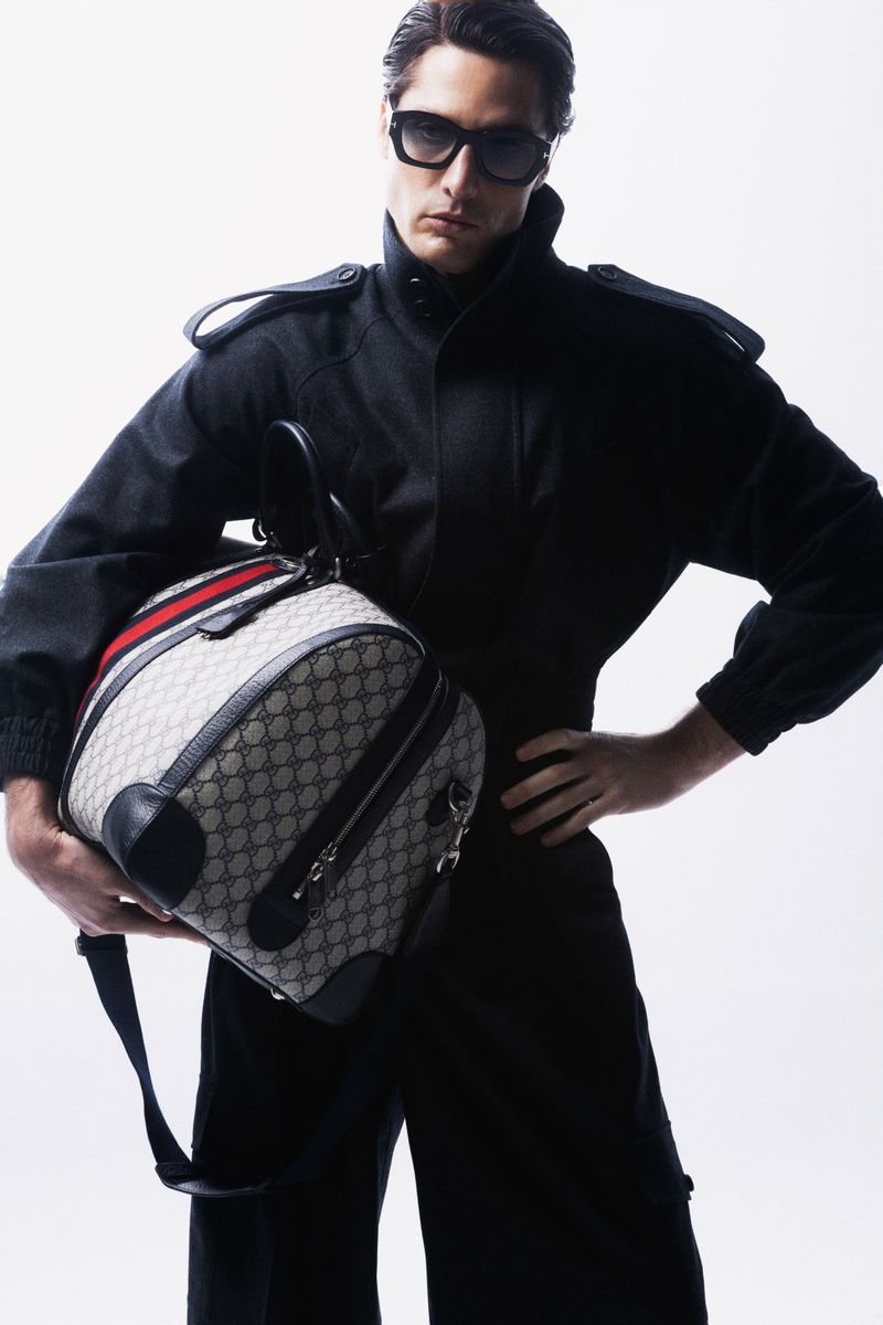 A man in a black jumpsuit and sunglasses holds a Gucci duffel bag in a studio setting, showcasing fashion and style.