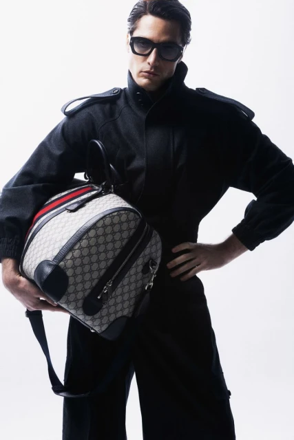 A man in a black jumpsuit and sunglasses holds a Gucci duffel bag in a studio setting, showcasing fashion and style.