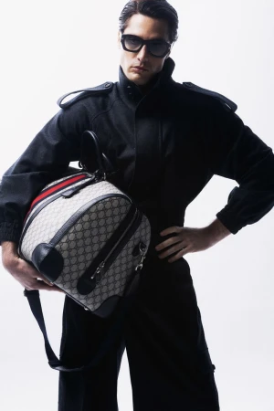 A man in a black jumpsuit and sunglasses holds a Gucci duffel bag in a studio setting, showcasing fashion and style.