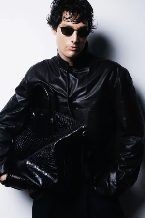 Portrait of an adult male model with curly dark hair, wearing sunglasses and a leather jacket, holding a textured leather bag.