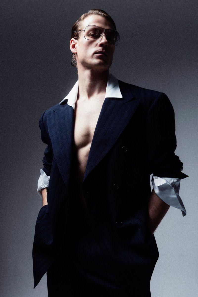 Fashionable adult male poses in an open, dark suit jacket with a white shirt collar and glasses, indoor in a photo studio.