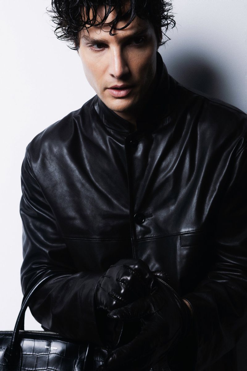 A brunette adult male poses in a leather jacket with a glossy leather bag in an eye-level portrait with a neutral expression.