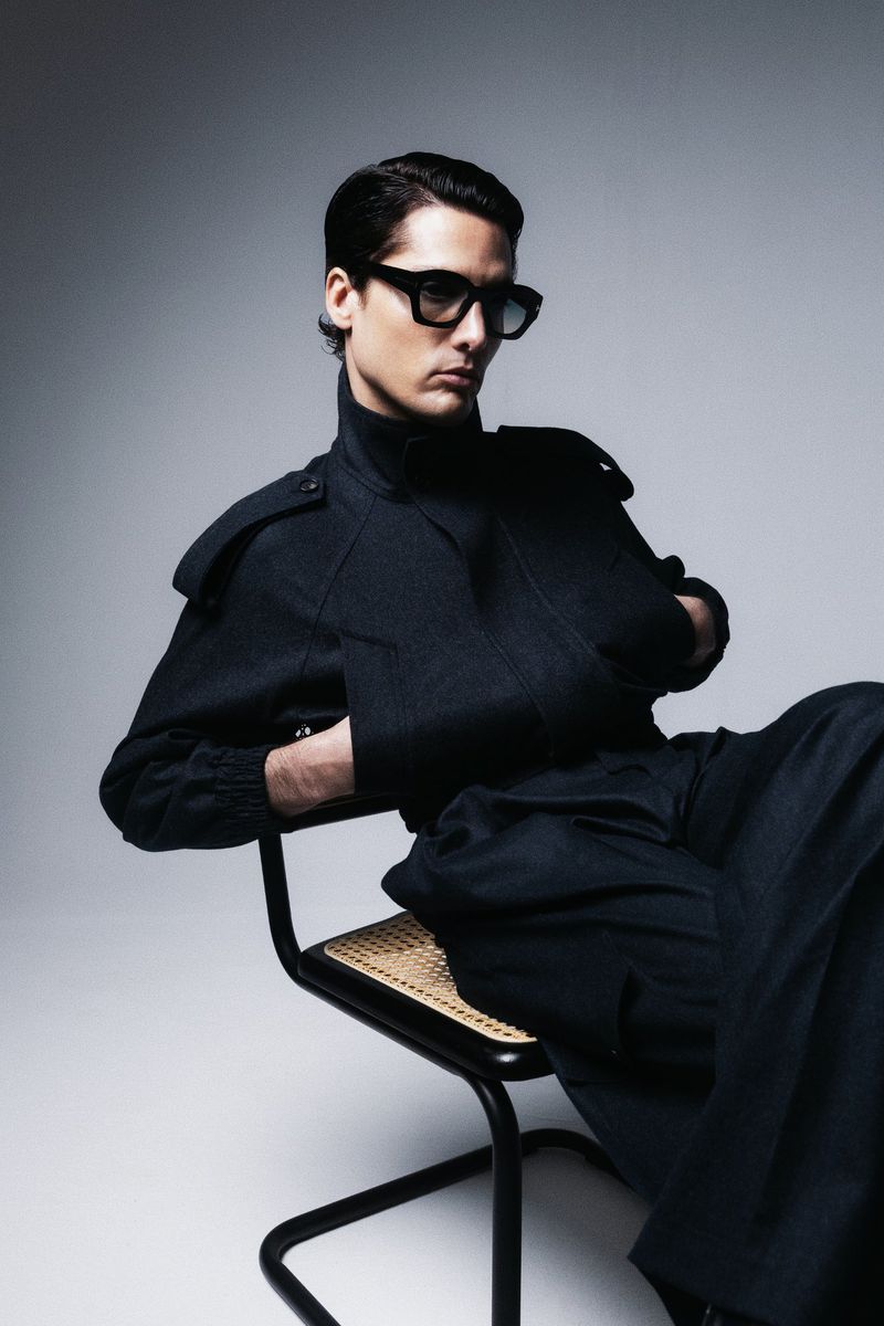 Fashionable adult male in stylish black jacket and pants sitting in a chair against a grey studio backdrop, wearing sunglasses.