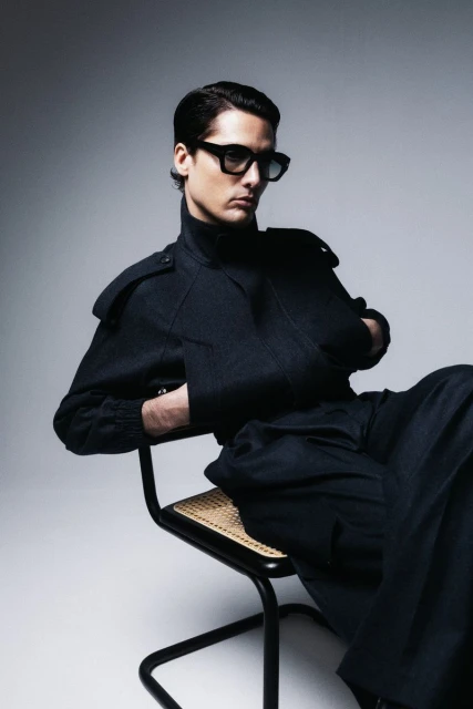 Fashionable adult male in stylish black jacket and pants sitting in a chair against a grey studio backdrop, wearing sunglasses.