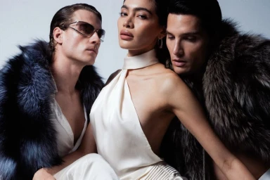 Model Priscilla Balanta in a cream halter top posed closely between two men in faux fur coats against a white studio background.