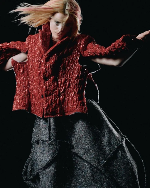 A young woman with blonde hair and a red textured jacket poses against a dark background wearing a grey patterned skirt. Fashion shot.