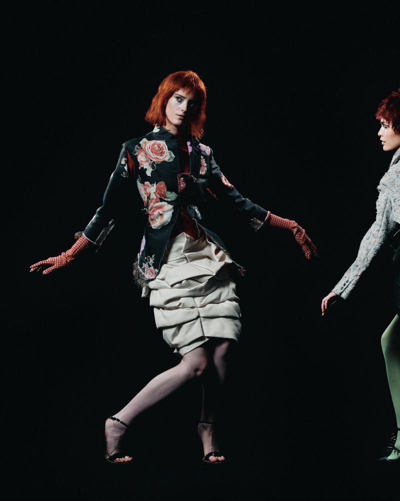 Two red-haired models pose in avant-garde fashion against a stark black background in a studio setting. The first model is dominant.