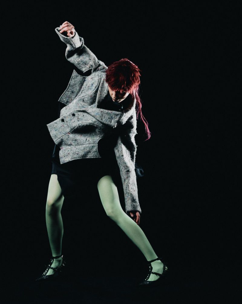 Fashionable person poses in grey tweed jacket, black dress, and light green tights against a black background in a studio shot.