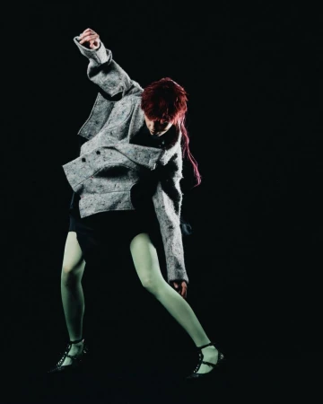 Fashionable person poses in grey tweed jacket, black dress, and light green tights against a black background in a studio shot.