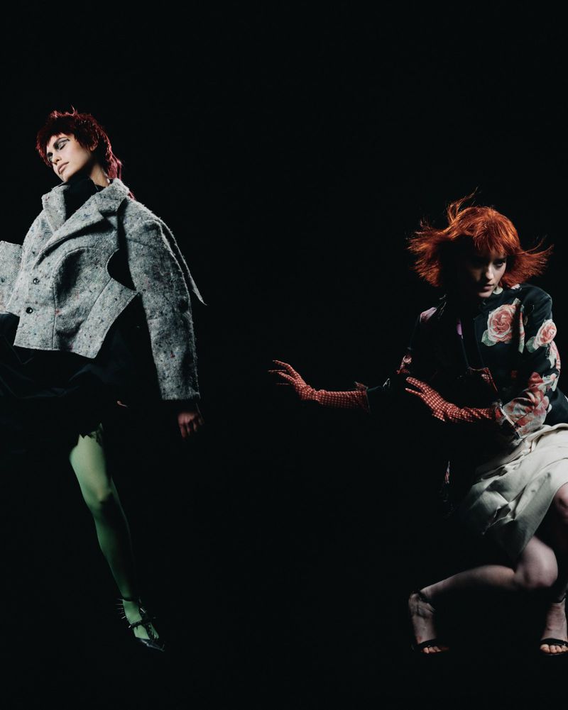 Two fashion models with red hair pose in a dark studio wearing stylish jackets, gloves, and dresses, showcasing modern editorial style and design.