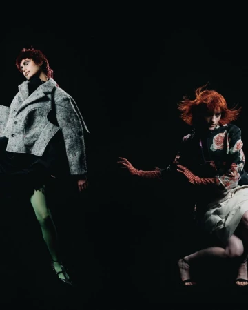 Two fashion models with red hair pose in a dark studio wearing stylish jackets, gloves, and dresses, showcasing modern editorial style and design.