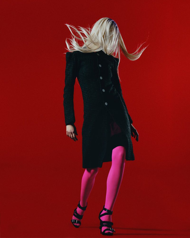 Woman in black coat with pink tights poses against red background, her blonde hair flowing. Fashion photography with bold colors.
