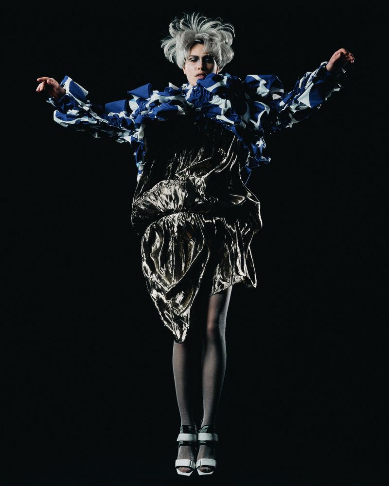 A person in a fashion editorial jumps in the air against a dark backdrop, wearing a blue and white patterned top and a gold skirt.