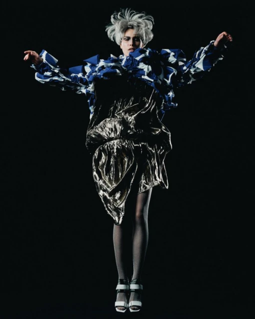 A person in a fashion editorial jumps in the air against a dark backdrop, wearing a blue and white patterned top and a gold skirt.