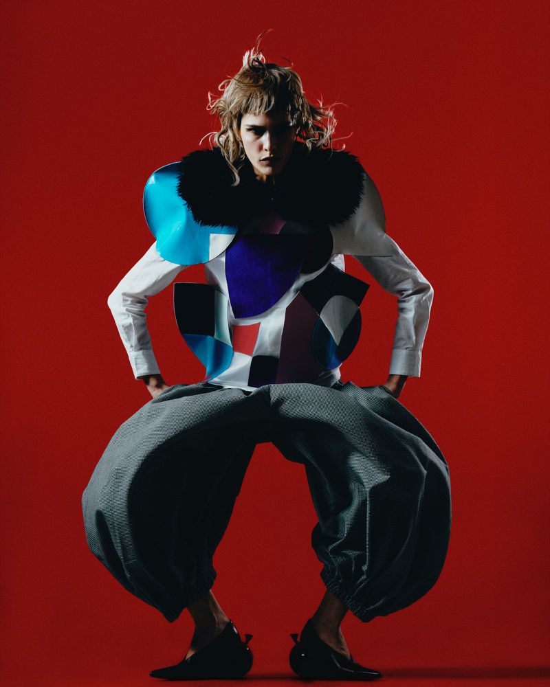 Avant-garde fashion portrait: model in sculptural top, voluminous grey trousers poses against a solid red background in studio lighting.