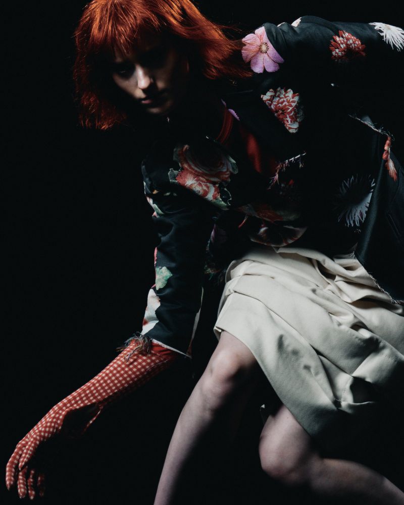 Woman with red hair wearing floral top and light skirt posing in a dark studio. Fashion editorial portrait with moody atmosphere.