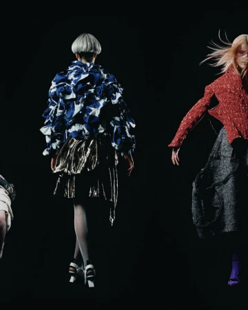 Fashion models walk the runway in avant-garde clothing on a black background. The details and textures of the fabrics are highlighted.