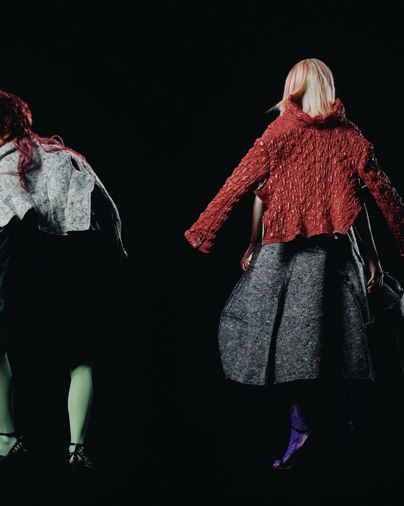 Two fashion models on a black backdrop wearing a bright red textured jacket, gray skirt, green tights, and black high heels.