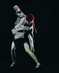 Model in a speckled grey oversized jacket and pale green tights in a dynamic pose against a solid black backdrop.