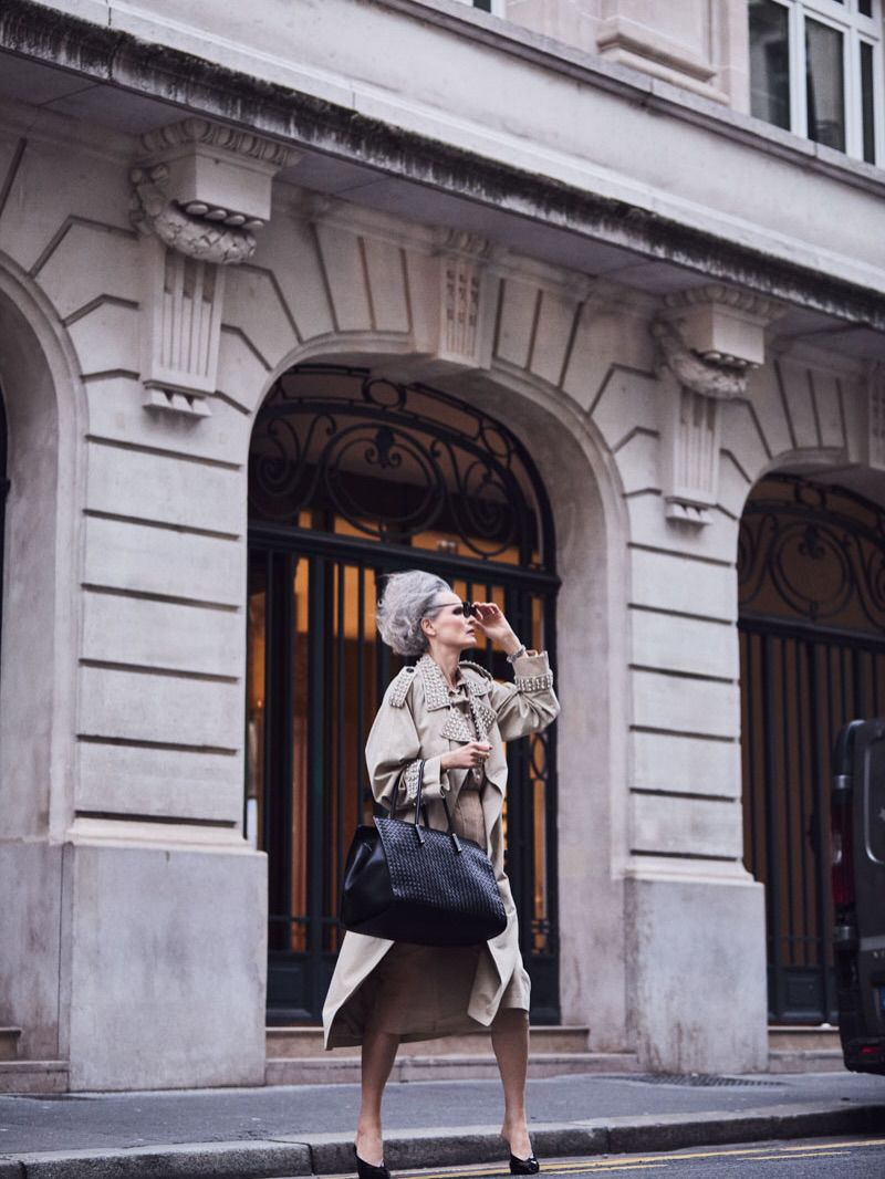 Mature woman with gray hair wearing a trench coat, sunglasses, and heels, standing in an urban setting, holding a handbag.
