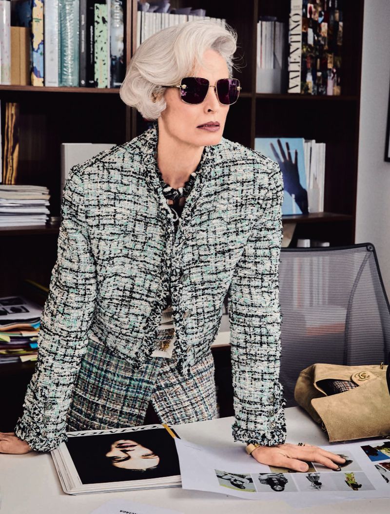 Mature woman with short grey hair wearing sunglasses and a tweed jacket in an office setting, seated behind a white desk with books.