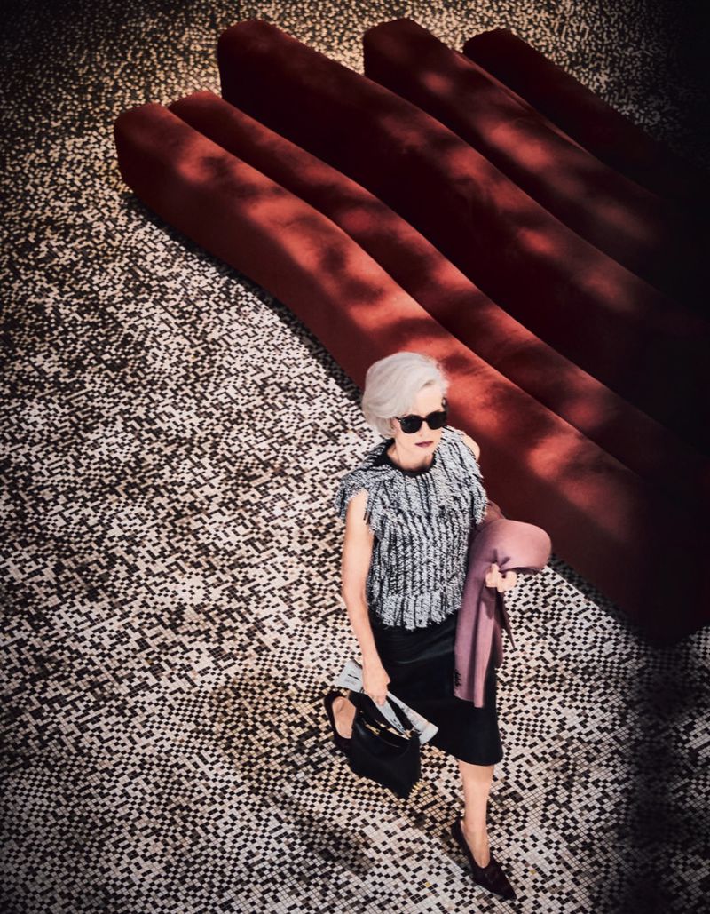 Elegant woman in stylish black-and-white tweed top, black skirt, and sunglasses walks on a patterned floor with red geometric shapes.