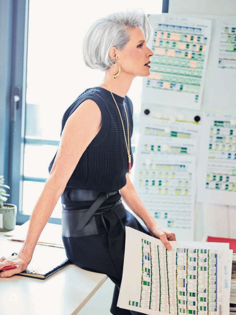 Elegant silver-haired woman in profile at bright office desk with color palettes, wearing sleeveless knit top and long gold pendant necklace.