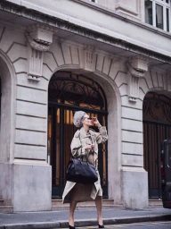 Sylke Golding in a beige trench coat and sunglasses carrying a large black handbag outside an ornate building.