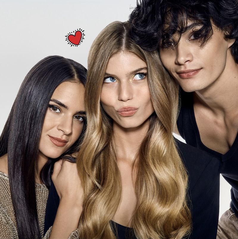 Three young adults pose closely together for a beauty advertisement. The blonde woman in the center looks off camera. A red heart is in the background.