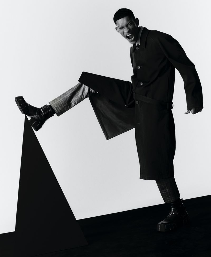 Monochromatic fashion shot of Black male model in trench coat and boots posing dramatically on a black geometric shape against white backdrop.