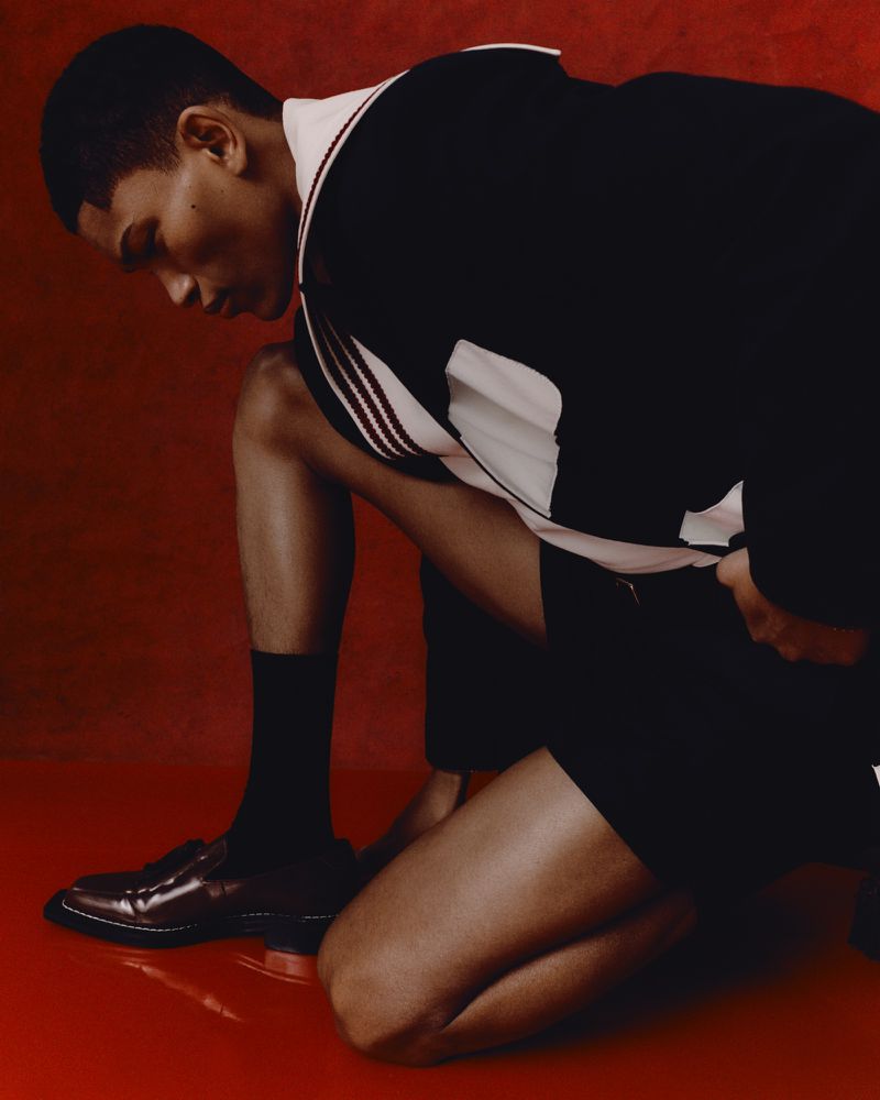Fashion shot of a Black male model kneeling in a black and white varsity jacket, black shorts and black socks in front of a red backdrop.