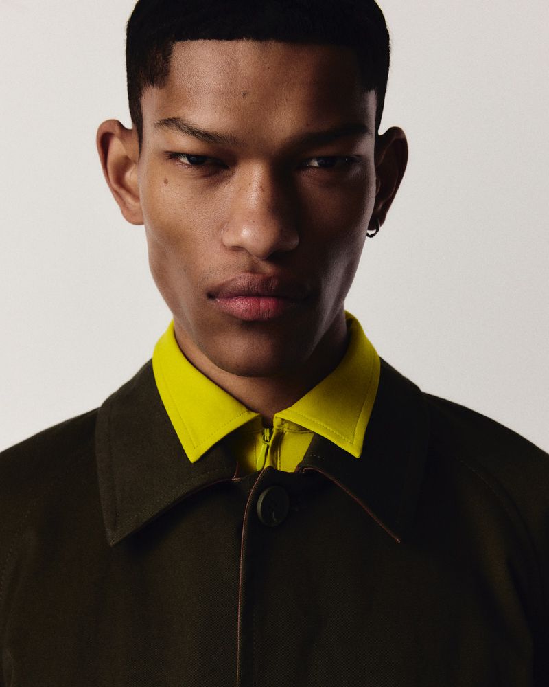 Portrait of a young Black man with short dark hair, wearing an olive coat with a yellow shirt, captured in a studio setting.