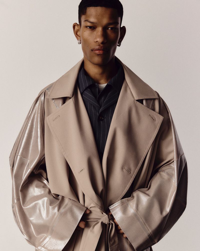A young Black male model poses in a tan trench coat and striped shirt against a neutral background, looking directly at the camera.
