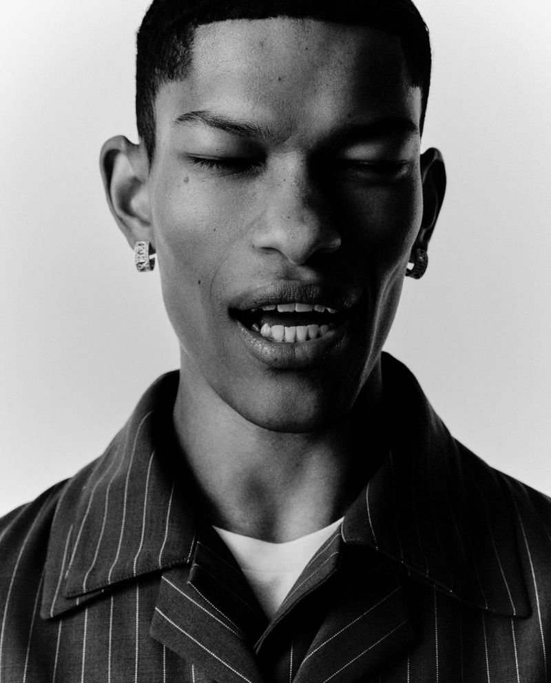 Portrait of a Black man with his eyes closed, wearing earrings and a striped button-down shirt in a black and white, eye-level studio shot.
